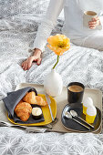 Croissants, eggs, coffee and yellow poppy on breakfast tray on bed with woman in background