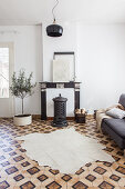 Tierfellteppich auf gemustertem Fliesenboden, graues Sofa, Holzofen vor stillgelegter Kamin und Olivenbäumchen im Wohnzimmer