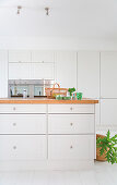 Kücheninsel mit weißen Schubladen und Holzplatte, im Hintergrund Einbauschrank mit integrierten Küchengeräten