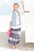 A young blonde woman on a beach wearing a white blouse, a denim gilet and a purple summer skirt