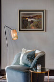 Armchair with scatter cushions between standard lamp and side table in front of painting on wall