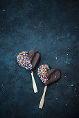 Heart-shaped vegan biscuits on sticks covered with dark chocolate and sprinkles