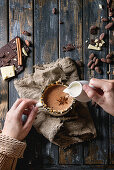 Female hands hold vintage mug of hot chocolate, decorated by nuts, caramel, spices, and pouring cream