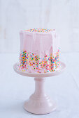 A pink frosted cake with colorful sprinkles on a pink stand