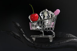 Schwarze Hand hält Mini-Einkaufswagen gefüllt mit Eiswürfeln und Kirsche