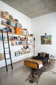 Wall-mounted shelves with library ladder next to couch on zebra-skin rug in bright interior with concrete ceiling and concrete floor