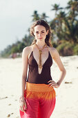 A young brunette woman on a beach wearing a brown bathing suit and harem trousers