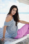 A young, long-haired woman on a lounger on a beach wearing a grey blouse and pink trousers