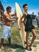 Junge Frau in schwarzem Abendkleid und junger Mann in Badeshorts mit Surfbrett
