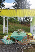 Tisch mit grüner Tischdecke, Stuhl, Baumstamm-Hocker und Sitzkissen unter Sonnensegel