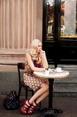 Junge blonde Frau in braunem Sommerkleid sitzt vor einem Café