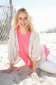 A young blonde woman sitting on a beach wearing a white blouse over a pink top