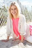 A young blonde woman sitting on a beach wearing a white blouse over a pink top