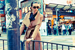 A young woman on a pavement wearing sunglasses and a camel-coloured coat