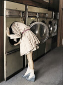 A young woman looking into a washing machine in a laundromat wearing a light trench coat