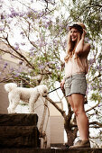 A young woman wearing a hat, a light top and shorts on a stone wall with a dog