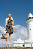 A blonde woman wearing a dark dress with a belt and a pale anorak by a water tower