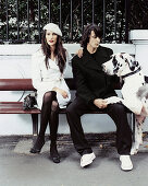 A dark-haired woman wearing a beret and a light trench coat with a man in a black outfit and a Dalmatian on a bench