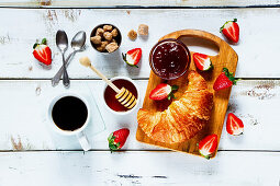 Rural breakfast with fresh croissant, cup of coffee, fruity jam, honey and strawberries on rustic white wooden background