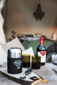 Cutlery, thermos flask, glasses and bottle of red wine on wooden table outside tent