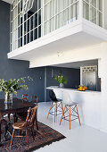 White kitchen counter and bar stools below gallery and dining table and chairs in high-ceilinged room