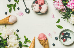 Pink strawberry and coconut ice cream scoops, sweet cones and peony flowers bouquet over white background