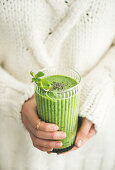 Matcha green vegan smoothie with chia seeds and mint in glass in hands of female wearing white sweater