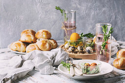 Easter table setting with colored orange eggs, hot cross buns, green branches decorated, empty white plate with cutlery, glass of lemonade drink