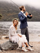 A bride wearing a white wedding dress sitting on a rock with a groom fishing at a lake