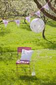 Romantic seat below lantern and bunting on flowering cherry tree:handmade cushion on chair next to branch wound with white yarn and decorated with crocheted flowers