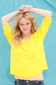 A young blonde woman wearing a yellow jumper and an apricot t-shirt against a blue background