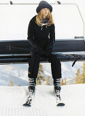 A blonde woman wearing a black ski suit on a ski lift with ski