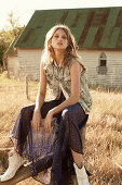 A young woman wearing a denim gilet and a long dress sitting on a fencepost