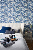 Tea service on tray on bed against wall covered in blue-and-white, leaf-patterned wallpaper