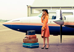 A brunette woman wearing an orange jumper and a matching skirt standing by a plane with suitcases