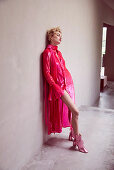 A blonde woman wearing a pink dress and matching ankle boots leaning on a wall
