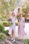 A blonde woman wearing a summer dress holding a bunch of flowers and a brunette woman wearing a delicate pink summer dress