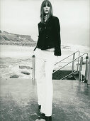 A young woman wearing a black jacket and white trousers by the sea