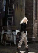 A blonde woman wearing a black blazer, jeans and high heel in front of an industrial building