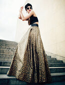 A young woman wearing a black turban, sunglasses, a crop-top and a leopard print maxi skirt