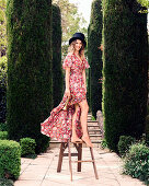 A young woman wearing a top hat and a summer dress standing on a step stool in a park