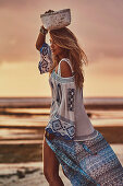 A blonde woman on a beach at sunset wearing an embroidered summer dress with basket on her head