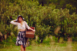 A blonde woman wearing a lace top and a floral skirt with an empty fruit crate in a garden