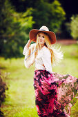 A blonde woman wearing a hat, a white top and a patterned skirt in a garden