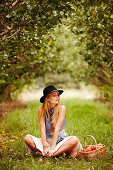 Blonde Frau mit Hut in weißem Kleid und Weste sitzt unter einem Apfelbaum