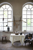 Bowls and collection of feathers on table, furry blanket on chair and stool below arched windows in loft apartment