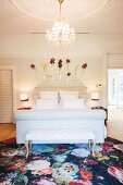 Elegant bedroom with white double bed, clothes bench, chandelier and flowers on the wall