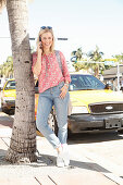 A blonde woman wearing jeans and a floral blouse on the phone outside