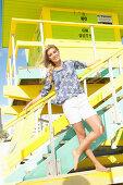 Blonde Frau in Shorts und geblümter Tunika steht an Strandhaus