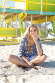 Blonde Frau in Shorts und geblümter Tunika sitzt vor Strandhaus
